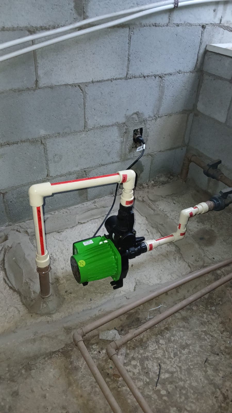 Bomba verde instalada em tubulação branca diante de uma parede de blocos.