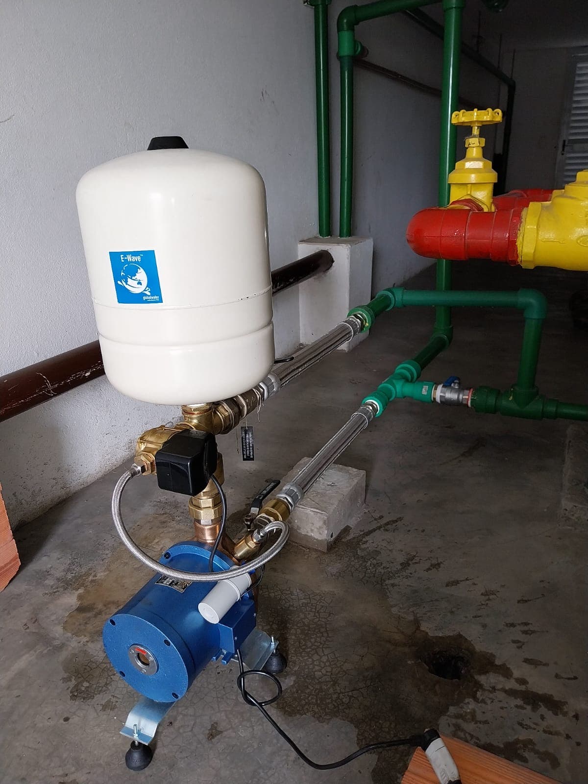 Bomba azul conectada a um vaso branco e a tubulações verdes por mangueiras metálicas.